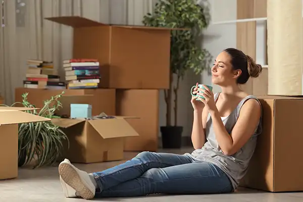 young woman sipping coffee and smiling happily near moving boxes with peace of mind for having a simplified small job move